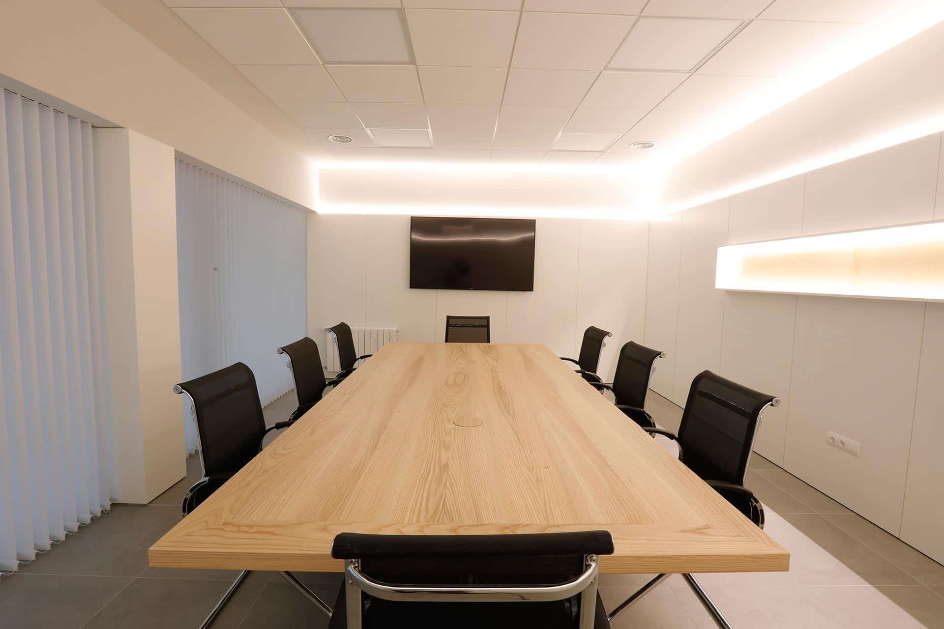 Sala de reuniones blanca con mesa de madera e iluminación indirecta en reforma de oficina de diseño diseñada por Moah Diseño Interior en Cantabria